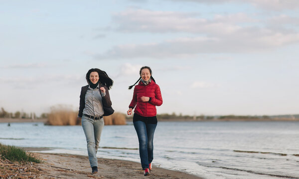 Couple Of Caucasian Girlfriends In Blue Jeans, A Jacket And A Shirt Run Along The River Bank In The Evening Of Sunset And Laugh With Joy. The Concept Of A Spring Walk Outdoors With Beloved Sister.