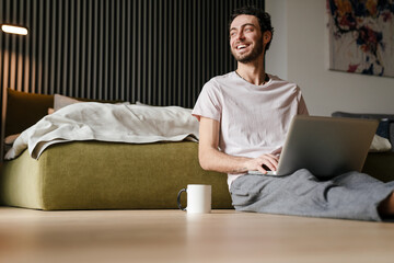 Handsome joyful man using laptop and laughing while sitting on floor
