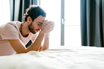 Focused unshaven man praying while holding hands together in bedroom