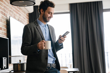 Handsome smiling guy using mobile phone while drinking coffee