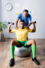 Fototapeta na wymiar african american sportsman training with dumbbells on fitness ball near physiotherapist during rehabilitation