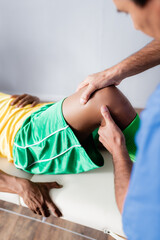 blurred middle aged masseur massaging knee of african american sportsman in sportswear in clinic - obrazy, fototapety, plakaty