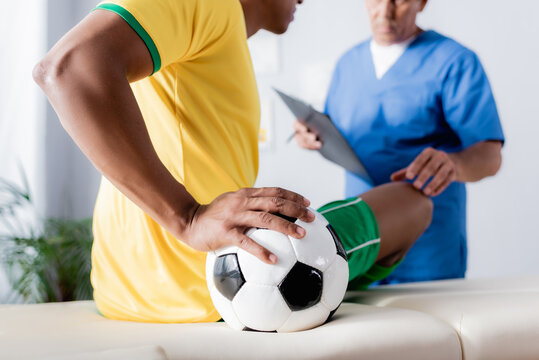 cropped view of injured african american football player sitting with ball on massage table near doctor with clipboard on blurred background