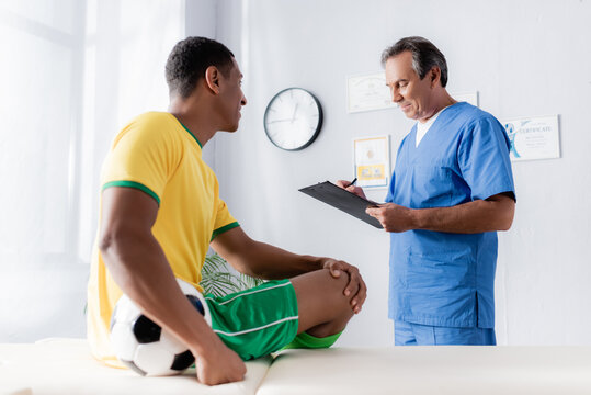 doctor writing prescription near smiling african american football player sitting with ball on massage table