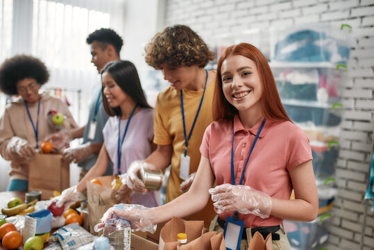 Young Female Volunteer Smiling At Camera While Packing Food And Drinks Donation Into Paper Bags And Box, Small Group Of People Working In Charitable Foundation