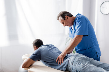 middle aged chiropractor working with young and injured african american patient on massage table in clinic