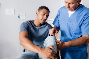 mature chiropractor pointing with finger at injured knee of african american man in clinic - Powered by Adobe