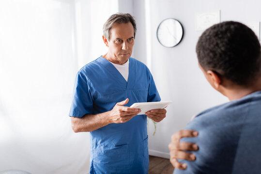 mature doctor holding digital tablet near african american patient on blurred foreground