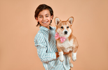 Cute smiling boy in a striped shirt holds a dog with a bow on his neck isolated on a powdery peach background.Valentine's Day.