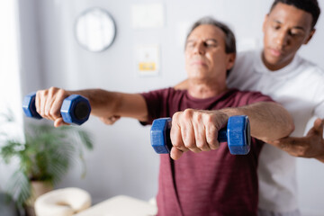 middle aged man exercising with dumbbells near african american physiotherapist on blurred background