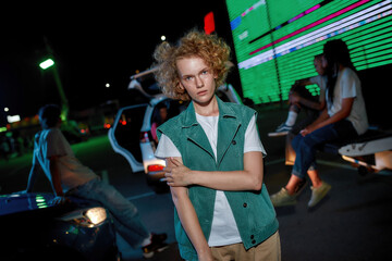 A well-dressed artistic redhead young woman with freckles looking into a camera standing outside with her left hand over her chest with a led screen behind