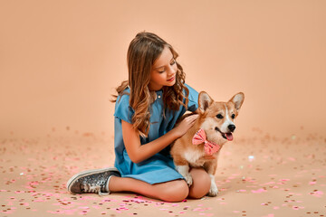 Cute pretty girl with curls sits on the floor with confetti and hugs a dog with a bow on the neck isolated on a powdery peach background. Valentine's Day.