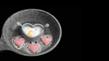 Cooking delicious breakfast with scrambled eggs and sausages in the shape of a heart in a gray frying pan isolated on black background, banner, copy space, top view. Valentine's Day Cooking