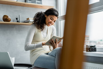 cheerful african american woman reading book while sitting near vintage camera on windowsill, blurred foreground