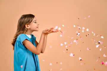 A cute girl in a blue dress and curly hair blows valentines hearts-shaped confetti from her palms...