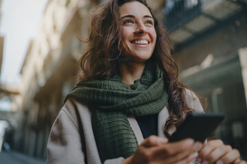 Young attractive smiling broadly woman wearing light-colored coat and green scarf, walking city streets on a sunny day and using smartphone for communication. Good vibes. Modern technology advantages