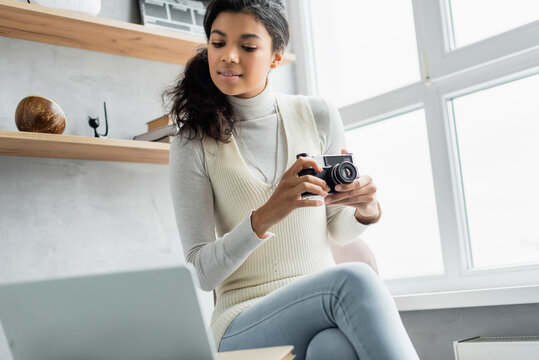 young african american woman holding vintage photo camera while sitting near laptop on blurred foreground