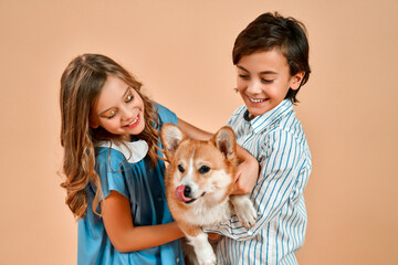 Cute pretty girl with curls in a blue dress and a boy in a shirt hug a funny dog ​​with a bow on his neck isolated on a powdery peach background.Valentine's Day.