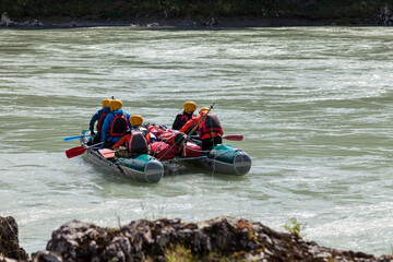 A team of people rafting in equipment, lifejackets and yellow helmets on a blue inflatable boat along a mountain river with rapids with raised oars.