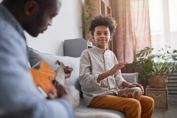 Mature African American man and his teen son spending great time together at home playing music on guitar and djembe