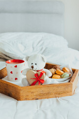 Valentine's day breakfast in bed for Lover. Teddy Bear with a heart-shaped gift box with red ribbon, a cup of coffee, and macaroons cookies on the wooden tray. Romantic morning. Vertical. Copy space
