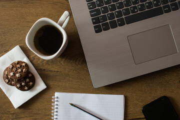 Notebook with pen, cookies, cup of coffee, smart phone and laptop on wooden desk, top view. Work from home concept