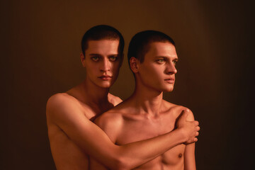 Fototapeta na wymiar Portrait of young half naked twin brothers posing together in studio, standing isolated over brown background, front view