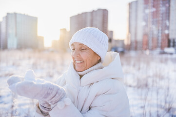 Happy elderly senior mature woman in white warm outwear playing with snow in sunny winter outdoors. Retired healthy people holiday vacation winter activities, active lifestyle concept.