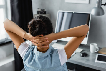 Fototapeta premium back view of african american freelancer sitting with hands behind head near computer on blurred background