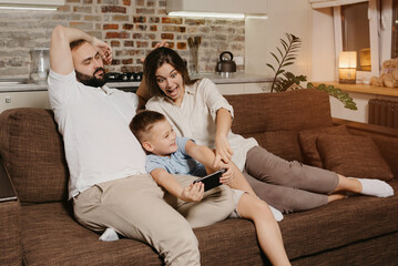 A dad, a son, and a mom on the sofa. A happy child is playing in the game on the smartphone near parents in the evening at cozy home. The mother is surprised.