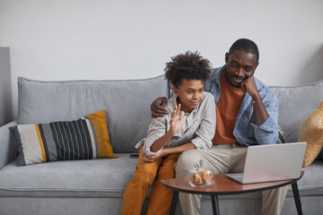 Modern father and son sitting on sofa in front of laptop screen starting video call with relatives...