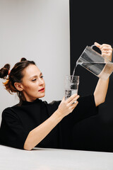 The girl in the black dress pours water from the carafe into a glass on a black and white background. High quality photo