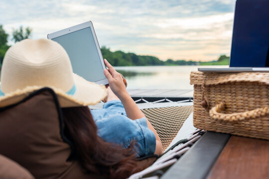 Woman with laptop and tablet remotely working on hammock and enjoying lake view. Remote place online work concept.