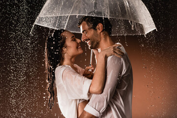 side view of happy couple embracing while standing with umbrella under rain on dark background - Powered by Adobe