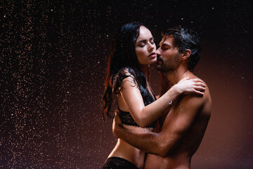 wet, muscular man hugging seductive woman in black lace lingerie under rain on dark background