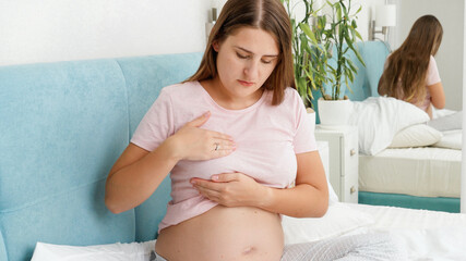 Young pregnant woman in pajamas sitting on bed and examining her breast for cancer. Concept of pregnancy healthcare and medical examination