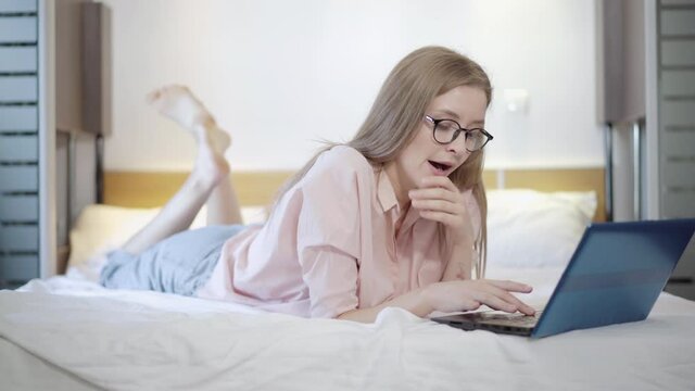 Exhausted young beautiful businesswoman in eyeglasses lying on bed and typing on laptop keyboard. Portrait of tired yawning Caucasian woman working online at home in bedroom. Freelance concept.