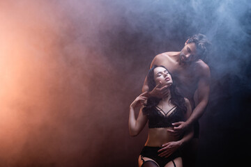 Fototapeta na wymiar shirtless muscular man embracing neck of sexy woman in black lace underwear on dark background with smoke