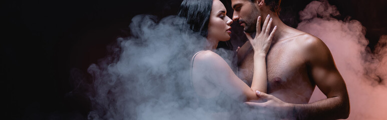 sexy woman touching shirtless man while standing face to face on black background with smoke, banner