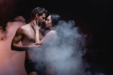 sensual woman touching sexy shirtless man while standing face to face on dark background with smoke