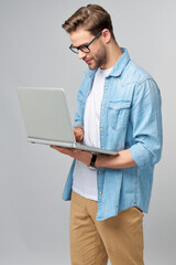 Concentrated young bearded man wearing glasses dressed in jeans shirt using laptop isolated over grey studio background
