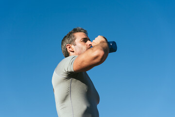 Mature fitness man with blue sky background, drinking from blue bottle. Copy space. Close up....