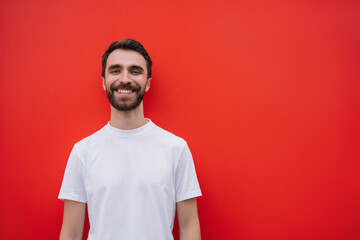 Portrait of stylish bearded man with happy emotional face looking at camera, smiling. Young handsome model wearing casual t-shirt isolated on red background 