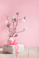 Beautiful white box with pink ribbon on the light wooden table with branches decorated with hearts