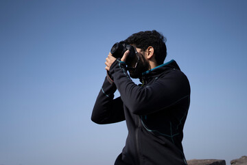Young indian boy clicking photos from his DSLR camera with blue sky in the background.