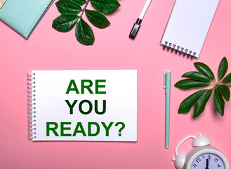 Question ARE YOUR READY is written in green on a white notepad on a pink background surrounded by notepads, pens, white alarm clock and green leaves. Educational concept