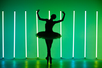 Beautiful young ballerina dancing classical ballet in a dark studio against a background of bright neon lights. Silhouette of a slender female dancer in a white tutu and pointe shoes.