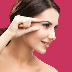 Skin care beauty concept. Portrait of smiling woman pointing brow or eye, touching skin, applying face cream, isolated on red color background. Brunette model at studio. Square composition.