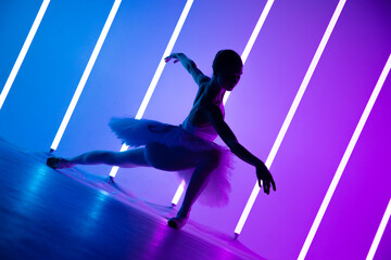 Young graceful ballerina in pointe shoes and a white tutu demonstrates her dancing skills. A beautiful classical ballet dancer against the backdrop of bright neon lights in a dark studio. Silhouette.