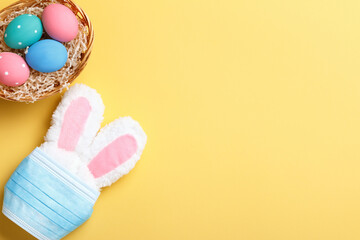 Easter concept. Rabbit in a protective medical mask and a basket with colored eggs on a yellow background.